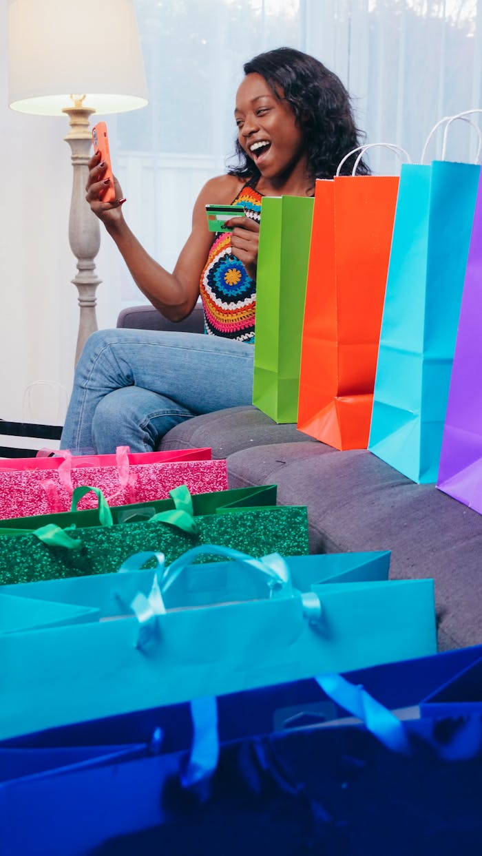 gallery-2 A woman enjoys online shopping with a smartphone, surrounded by colorful shopping bags indoors.